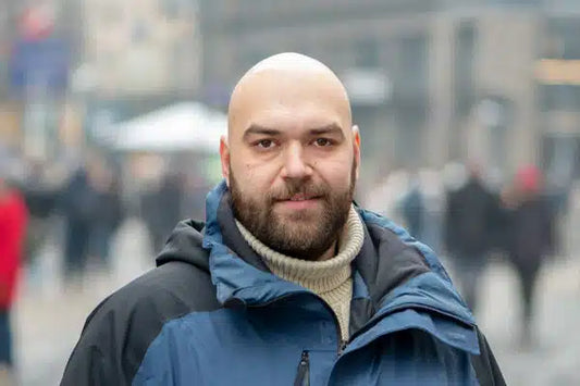 A bald man with a beard wearing a heavy winter jacket and turtleneck sweater standing outdoors in a city setting