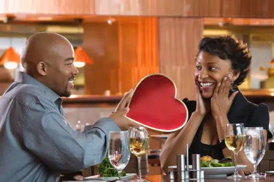 A smiling bald man with a shiny scalp giving a heart-shaped gift box to a surprised woman at a dinner table