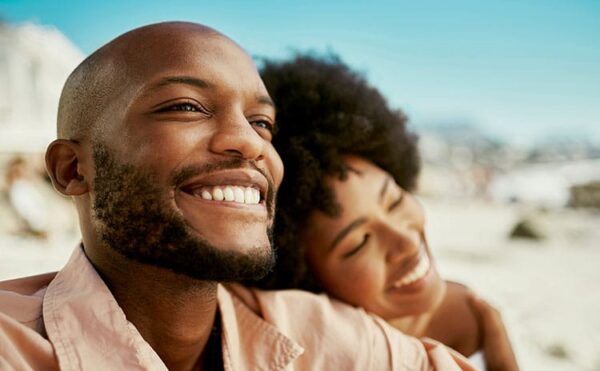 A close-up of a smiling bald man with a beard, with a woman resting her head on his shoulder as they both laugh together on a bright, sunny beach